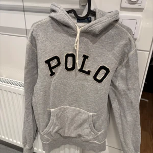 Grå hoodie från Ralph Lauren - Säljer en grå hoodie från Ralph Lauren med stor 'POLO' text på framsidan. Tröjan har en klassisk ficka och justerbar huva med snören. Perfekt för en avslappnad stil. Pris går att diskuteras