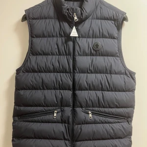 Mörkblå dunväst från Moncler - Snygg mörkblå dunväst från Moncler med dragkedja och två fickor framtill. Västen har en elegant design och Moncler-logga i svart på bröstet. Perfekt för lite svalare dagar. Västen kommer direkt från Moncler butiken i Barcelona, (2024 modell). Den är i storleken medium (3).