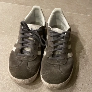 Adidas Gazelle grå - Snygga grå Adidas Gazelle sneakers med vita detaljer och klassiska tre ränder på sidan. Skorna har snörning och en bekväm passform. Perfekta för en avslappnad stil.