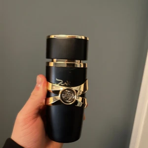 Asad Parfym från Lattafa - Elegant parfymflaska i svart med gyllene detaljer från Lattafa. Flaskan har en unik design med ett dekorativt band och arabiska bokstäver. Perfekt för den som söker en lyxig doftupplevelse.