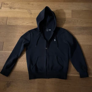 Ralph Lauren hoodie - Säljer en stilren från Ralph Lauren med dragkedja och klassisk logga på bröstet. Perfekt för en avslappnad look. 7/10 skick och kostar runt 2400 kr orginalpris.