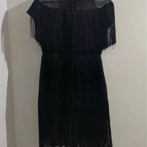 Svart fransklänning - Snygg svart klänning med fransdetaljer som ger en cool och trendig look. Perfekt för att sticka ut på festen. Klänningen har en offshoulder-design som ger en elegant touch.