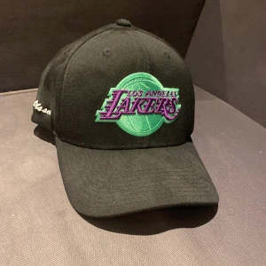 Svart keps från Mitchell & Ness - Snygg svart keps med Los Angeles Lakers-logga i lila och grönt framtill. Märket Mitchell & Ness är broderat på sidan. Perfekt för sportfantaster och streetwear-entusiaster. Text med ”Olsson” på sidan, går enkelt att sprätta bort men därav priset