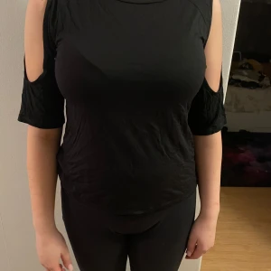 Svart topp med öppna axlar - Snygg svart topp med öppna axlar och korta ärmar. Perfekt för en stilren look. Toppen har en bekväm passform och är tillverkad i ett mjukt material.