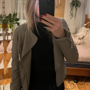 Grå skinnjacka med dragkedjor - Snygg grå skinnjacka med coola dragkedjor och bältesdetaljer i midjan. Perfekt för en stilren look. Jackan har långa ärmar och en asymmetrisk dragkedja framtill. Är figursydd vilket gör att den sitter väldigt bra i midjan.