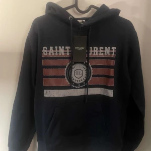 Mörkblå hoodie från Saint Laurent - Säljer en snygg mörkblå hoodie från Saint Laurent med tryck i rött och vitt. Hoodien har en klassisk design med dragsko i huvan och en stor ficka framtill. Perfekt för en avslappnad stil. 