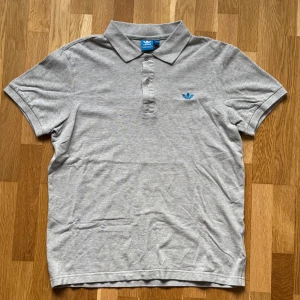 Adidas Grå Piké - Grå Piké med blå logga från Adidas.  Storlek: L, Armhåla till Armhåla: 52 cm, Längd: 66 cm, Axel till Armöppning: 19.5 cm.  Material: 90% Cotton 10% Polyester