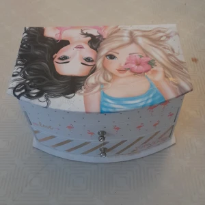 Smyckeskrin från TOPModel - Charmigt smyckeskrin från TOPModel med ett färgglatt motiv av två flickor på locket. Skrinet har en spegel inuti och flera fack för förvaring. Utsidan är dekorerad med flamingos och hjärtan i rosa och guld. Perfekt för att organisera dina smycken och småsaker.