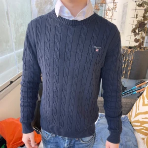 Mörkblå kabelstickad tröja från GANT - Säljer en stilren mörkblå kabelstickad tröja från GANT. Tröjan har en klassisk design med ribbade muddar och rund hals. Perfekt för en avslappnad men ändå snygg look. Passar bra till jeans eller chinos. Använd endast en gång.