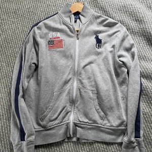 Grå tröja från Ralph Lauren - Säljer en grå tröja från Ralph Lauren med dragkedja och broderad logga på bröstet. Jackan har långa ärmar med mörkblå rand och en amerikansk flagga broderad på framsidan. 