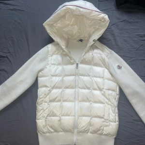 Vit Moncler Cardigan med huva - Säljer en stilren vit quiltad jacka från Moncler med huva och dragkedja. Jackan har långa ärmar och en mjuk, ribbad nederkant. Perfekt för kyligare dagar men även under våren. Jackan är dock like missfärgad pågrund av användning och dragkedjan saknar två bitar men den går upp och ner .