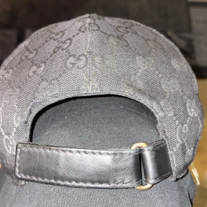 Svart keps med monogrammönster - Snygg svart keps med diskret monogrammönster och justerbart bälte i skinn. Har en grön och röd randdetalj på sidan. Perfekt för en stilren look. Jag säljer kepsen eftersom jag har bytt stil helt och inte har användning till den längre.