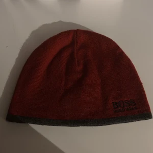 Röd mössa från Hugo Boss - Snygg röd mössa från Hugo Boss med en diskret logga i svart. Mössan har en grå kant och är perfekt för kyliga dagar. Den är tillverkad i ett mjukt material som håller dig varm och bekväm.