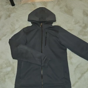 Grå huvtröja från Sail Racing, strl 176 - Säljer en grå huvtröja från Sail Racing med dragkedja framtill. Tröjan är tillverkad i en blandning av bomull och polyester, vilket gör den både bekväm och hållbar. Perfekt för kyliga dagar med sin långa ärmar och praktiska huva.