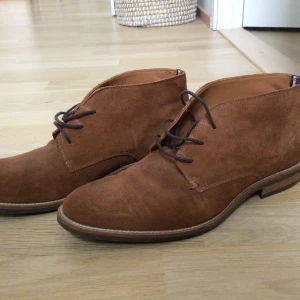 Bruna mockaboots från Tommy Hilfiger - Snygga bruna boots i mocka från Tommy Hilfiger med klassisk snörning. Perfekta för en stilren look. Skorna har en diskret logga på hälen och en robust sula för extra komfort.