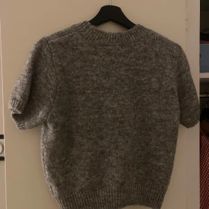 Grå stickad tröja - Mysig grå stickad tröja med korta ärmar. Perfekt för kyliga dagar och ger en avslappnad look. Ribbstickad vid ärmslut och nederkant för extra komfort.
