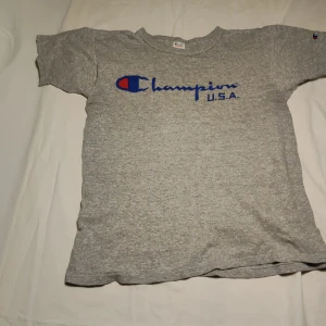 Grå t-shirt från Champion - Säljer en klassisk grå t-shirt från Champion med blå och röd logga på framsidan. T-shirten är kortärmad och tillverkad i en mjuk bomullsblandning. Perfekt för en avslappnad stil.