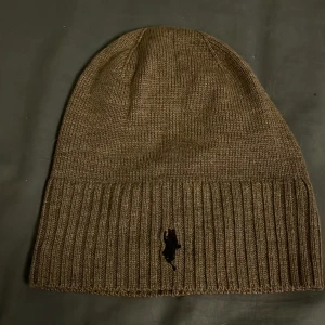 Brun stickad mössa från Polo Ralph Lauren - Grå stickad mössa från Polo Ralph Lauren i merinoull. Mössan har en ribbad kant och en broderad logotyp framtill. Pris kan diskuteras.