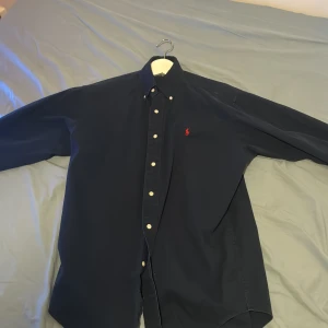 Mörkblå skjorta från Ralph Lauren - Säljer en stilren mörkblå skjorta från Ralph Lauren i 100% bomull. Skjortan har långa ärmar och knappar framtill samt en broderad logotyp i rött på bröstet. Perfekt för en klassisk look. Väldigt bra skick 10/10 perfekt till sommaren!