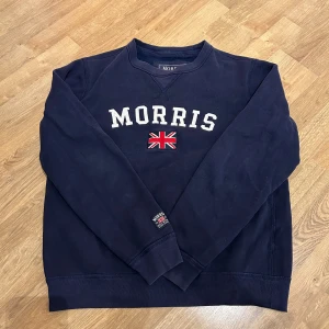 Morris tröja - Säljer en snygg mörkblå sweatshirt från Morris med text och brittisk flagga på bröstet. Tröjan har långa ärmar och rund halsringning. Perfekt för en avslappnad stil.