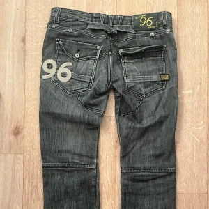 Svarta jeans från G-Star RAW - Snygga svarta jeans från G-Star RAW, modell Elwood 96. De har en unik design med broderad text och nummerdetaljer på baksidan. Perfekta för en cool och avslappnad stil. En knapp i bakfickan saknas. 