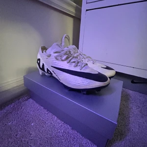 Vita och svarta fotbollsskor från Nike - Säljer ett par snygga vita och svarta fotbollsskor från Nike. De har en cool design med snörning och är perfekta för fotbollsträning eller match. Skorna ser ut att vara i bra skick och är redo för nya äventyr på planen.