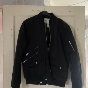 Svart bomberjacka - Säljer en svart bomberjacka i storlek M. Den har en klassisk design med dragkedja framtill och fickor med knappar. Perfekt för höst och vår!