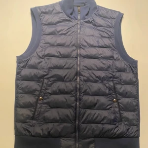 Blå quiltad dunväst från Polo Ralph Lauren - Säljer en snygg blå quiltad dunväst från Polo Ralph Lauren. Västen har en dragkedja framtill och två fickor med knappar. Perfekt för höst och vår när du vill hålla dig varm men ändå se stilren ut. Märkets ikoniska logga är broderad på bröstet. Passar perfekt för en casual look!