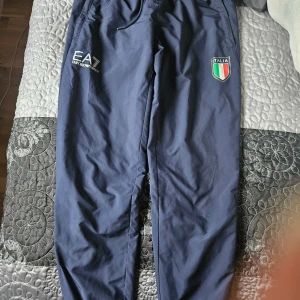 Blå träningsbyxor från EA7 Emporio Armani - Säljer ett par snygga blå träningsbyxor från EA7 Emporio Armani. De har en elastisk midja med dragsko och ett coolt Italienskt emblem på ena benet. Perfekta för träning eller en avslappnad dag. Materialet känns lätt och bekvämt.