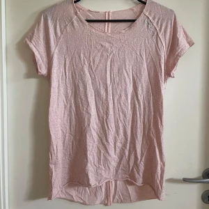 Ljusrosa kortärmad topp - Säljer en ljusrosa kortärmad yoga topp i ett mjukt och bekvämt material. Toppen har en rund halsringning och detalj med hål som löper längs ryggen. Perfekt för en avslappnad träningslook!