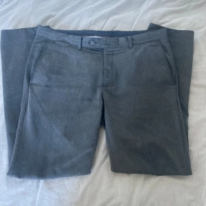 Grå kostymbyxor - Säljer ett par stiliga grå kostymbyxor W34 L32. Byxorna har en klassisk design med dragkedja och knappar framtill. De har också två bakfickor med knappar och bälteshällor för en perfekt passform. Straight fit och i bra skick