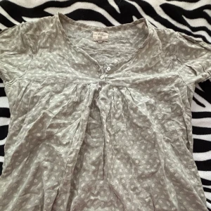 Grå mönstrad blus från Noa Noa - Säljer en söt grå blus med ett diskret mönster. Blusen har korta ärmar och en v-ringad hals med knappar. Perfekt för en avslappnad vardagslook!