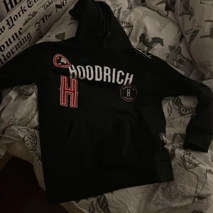 Svart hoodie från Hoodrich - Säljer en svart hoodie från Hoodrich med flera tryck och broderade detaljer i vitt och rött. Den har en klassisk känguruficka och justerbar huva. Perfekt för en avslappnad stil.