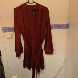 Vinröd långärmad klänning med knytband - Elegant vinröd klänning med långa ärmar och ett knytband i midjan. Perfekt för en stilren look. Tillverkad i ett mjukt och glansigt material som ger en lyxig känsla.