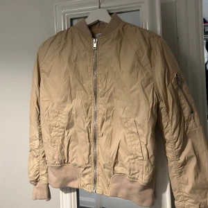 Beige Rebecca Stella bomberjacka  - Snygg beige bomberjacka med dragkedja och ribbade muddar. Jackan har ett coolt tryck med bokstäverna 'RS' förkortning för Rebecca Stella på ryggen. Perfekt för en avslappnad stil.