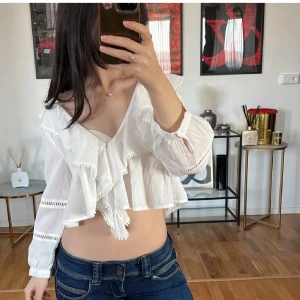 Vit blus med volanger - Säljer en söt vit blus med volanger och v-ringning. Blusen är croppad och har långa ärmar med spetsdetaljer. Perfekt för en somrig look!