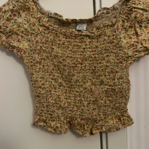 Blommig offshoulder topp - Charmig blommig offshoulder topp i gul och rosa med volangdetaljer. Perfekt för sommarens varma dagar. Den har en smockad design som ger en bekväm och flexibel passform.