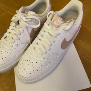 Nike sneakers i vitt och rosa - Snygga Nike sneakers i vitt med rosa detaljer. Skorna har klassisk snörning och en bekväm passform. Perfekta för en stilren look med en touch av färg. Använt max 2 gånger