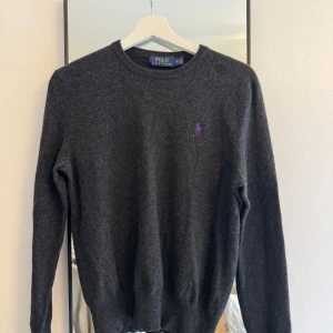 Mörkgrå tröja från Ralph Lauren - Säljer en stilren mörkgrå tröja från Ralph Lauren med rund hals och lila logga på bröstet. Tröjan är långärmad och har ribbade muddar vid ärmslut och nederkant. Perfekt för kyligare dagar.
