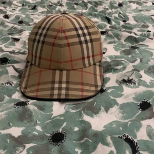 Rutig keps från Burberry - Snygg rutig keps från Burberry i beige, svart och röd. Kepsen har ett klassiskt rutmönster och justerbart spänne baktill för perfekt passform. Perfekt för att ge din outfit en stilren touch. 