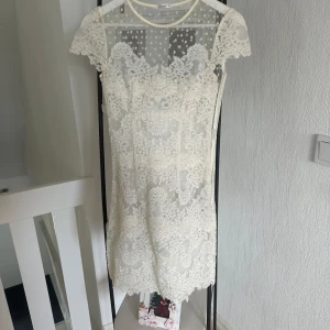 Vit spetsklänning från Ida Sjöstedt - Elegant vit spetsklänning från Ida Sjöstedt med korta ärmar och ett vackert blommigt mönster. Perfekt för speciella tillfällen med sin romantiska design och fina detaljer. Klänningen har en kort längd och en feminin passform.