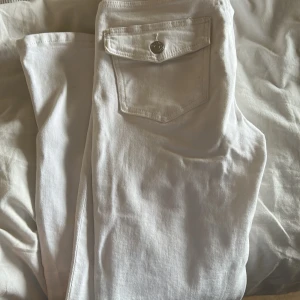 Vita jeansbyxor med fickdetalj - Snygga vita jeansbyxor med en unik fickdetalj på baksidan. Byxorna har en klassisk design med knappstängning på fickan, vilket ger en stilren look. Perfekta för en fräsch och modern stil.