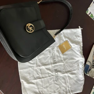 Svart axelväska från Michael Kors - Snygg svart axelväska från Michael Kors med ett stort guldfärgat MK-emblem på framsidan. Väskan har en stilren design med ett brett axelband och gott om plats inuti. Perfekt för att bära med sig det nödvändigaste med stil.