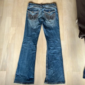 Blå bootcut jeans med broderade fickor - Snygga blå bootcut jeans med unika broderade detaljer på bakfickorna. Jeansen har en klassisk femficksdesign och en knappgylf. Perfekta för en avslappnad stil med en touch av elegans.