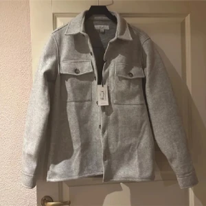 Grå overshirt från Erla of Sweden - Säljer en stilren grå overshirt från Erla of Sweden. Jackan har långa ärmar och knappar framtill samt två bröstfickor med knappar. Perfekt för lager-på-lager-stil under svalare dagar.