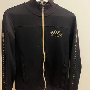 Svart tröja med dragkedja från Hugo Boss - Snygg svart tröja från Hugo Boss med gulddetaljer och dragkedja framtill. Tröjan har långa ärmar och ett stilrent tryck med BOSS-loggan på bröstet. Perfekt för en casual look.