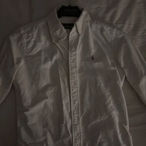 Vit skjorta från Ralph Lauren - Klassisk vit skjorta från Ralph Lauren med broderad logotyp på bröstet. Skjortan har en stilren design med knappar framtill och en krage som ger en tidlös look. Perfekt för en elegant och avslappnad stil.