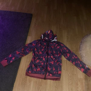 Amazon bape hoodie  - Säljer en cool hoodie med röd och svart camouflage och hajmotiv på huvan. Jackan har dragkedja och långa ärmar. Perfekt för en unik och trendig stil. Aldrig använt 