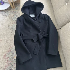 Svart kappa från Hollies - Snygg svart kappa från Hollies med huva och knytbälte i midjan. Den har en stilren design med knappar framtill och är perfekt för kyligare dagar. Kappan är tillverkad i ett material utan animaliska produkter.