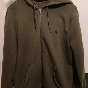 Olivgrön hoodie från Ralph Lauren - Säljer en olivgrön hoodie från Ralph Lauren med dragkedja och huva. Tröjan har en broderad logga på bröstet och praktiska fickor framtill. Perfekt för en avslappnad stil.